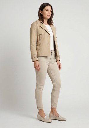 Kvinde stående, iført en beige kort jakke, hvid top, beige slanke bukser og beige slip-on sko med lave hæle.