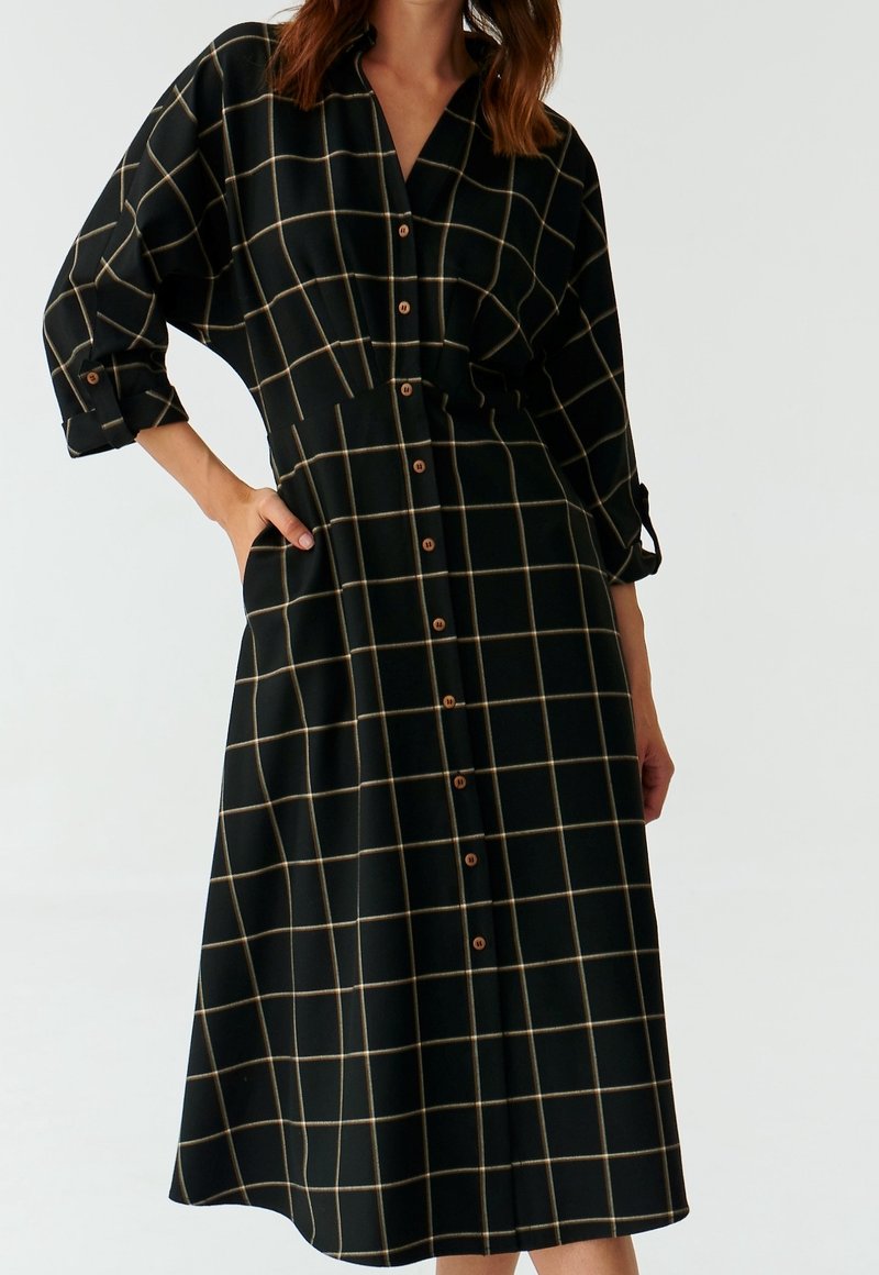 Femme portant une robe midi noire avec un motif quadrillé beige, devant boutonné, manches longues retroussées et une main dans la poche.