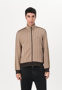 Hnedá patterned zipsová bunda s vysokým golierom, rebrovanými manžetami a lemom, so zeleným logom na hrudi a kontrastnými hnedými bočnými pruhmi.