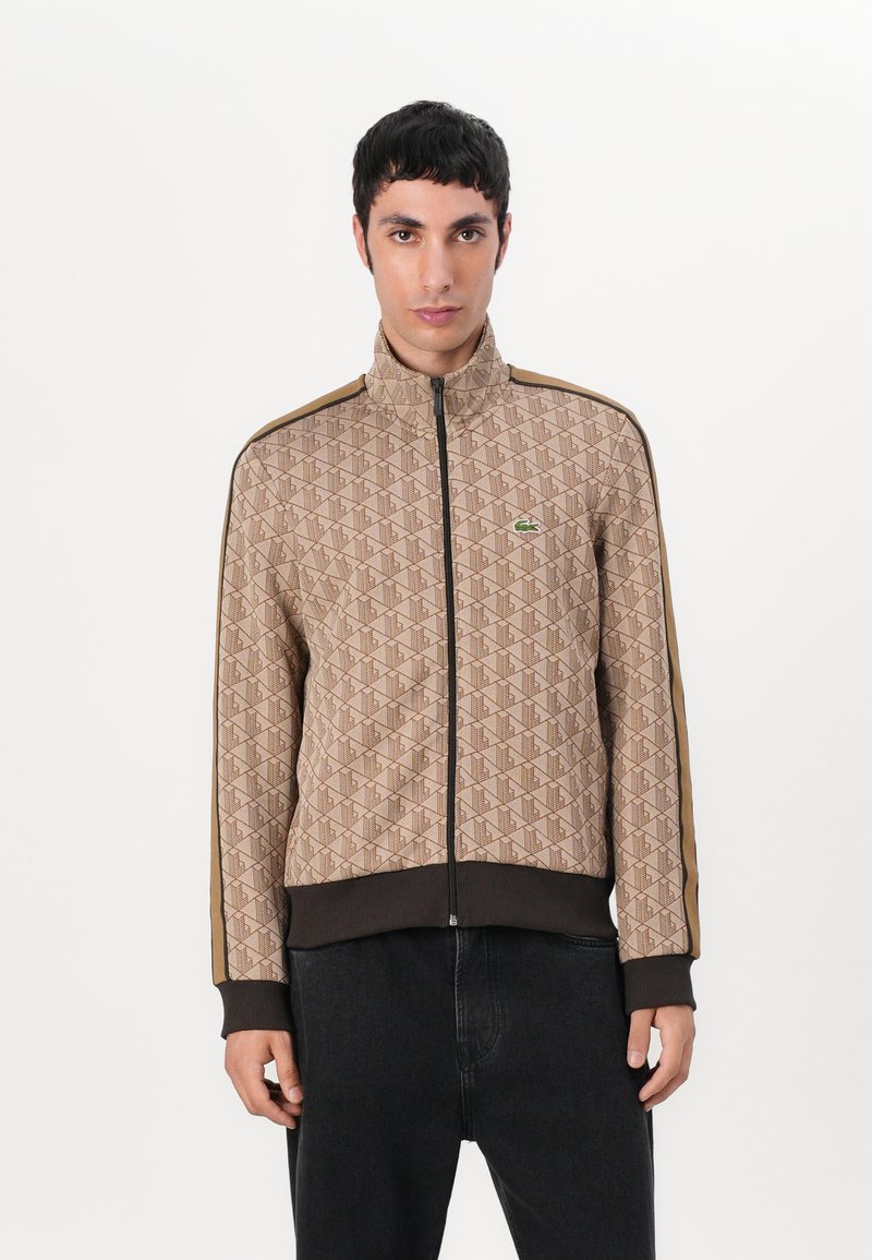 Hnedá patterned zipsová bunda s vysokým golierom, rebrovanými manžetami a lemom, so zeleným logom na hrudi a kontrastnými hnedými bočnými pruhmi.