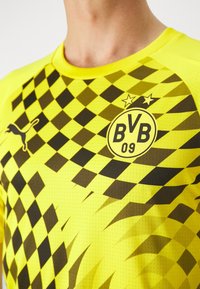 Camiseta deportiva amarilla con un patrón de tablero de ajedrez en negro. Incluye un logo redondo con "BVB 09" y dos estrellas encima. Logo de Puma en el hombro.