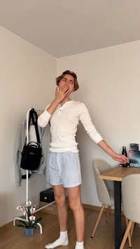 Jaunas žmogus, vilkintis baltu ilgomis rankovėmis marškiniais ir dryžuotomis šortais, �žiovauja ir tempiasi minimalistinėje patalpoje su drabužių stovu ir stalu.