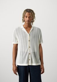 Camisa de punto ligera de manga corta en blanco, con cuello tipo polo y un cierre de botones al frente con botones en tono beige y un patrón texturizado.