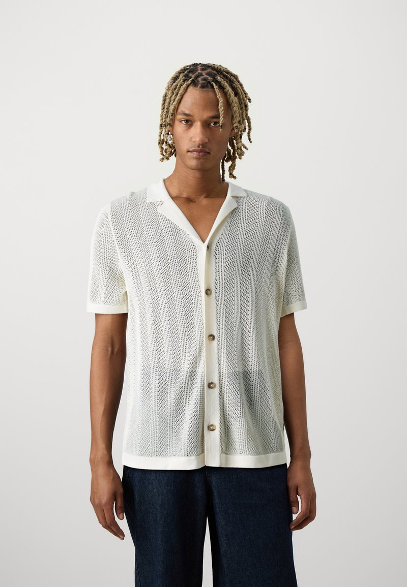 Camisa de punto ligera de manga corta en blanco, con cuello tipo polo y un cierre de botones al frente con botones en tono beige y un patrón texturizado.