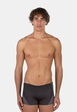 Giovane uomo con capelli castani ricci in piedi di fronte, indossa slip da bagno neri aderenti con dettagli chiari a fantasia, su uno sfondo bianco semplice.