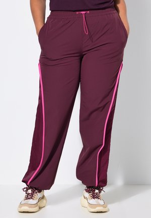 Pantalones de chándal granate con franjas laterales rosas, combinados con zapatillas chunky beige y blancas, manos en los bolsillos, de pie contra un fondo liso.