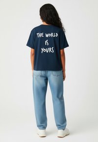 T-shirt bleu marine à manches courtes avec un texte blanc au dos disant "LE MONDE EST À VOUS"; associé à un jean léger et ample de couleur bleu clair.