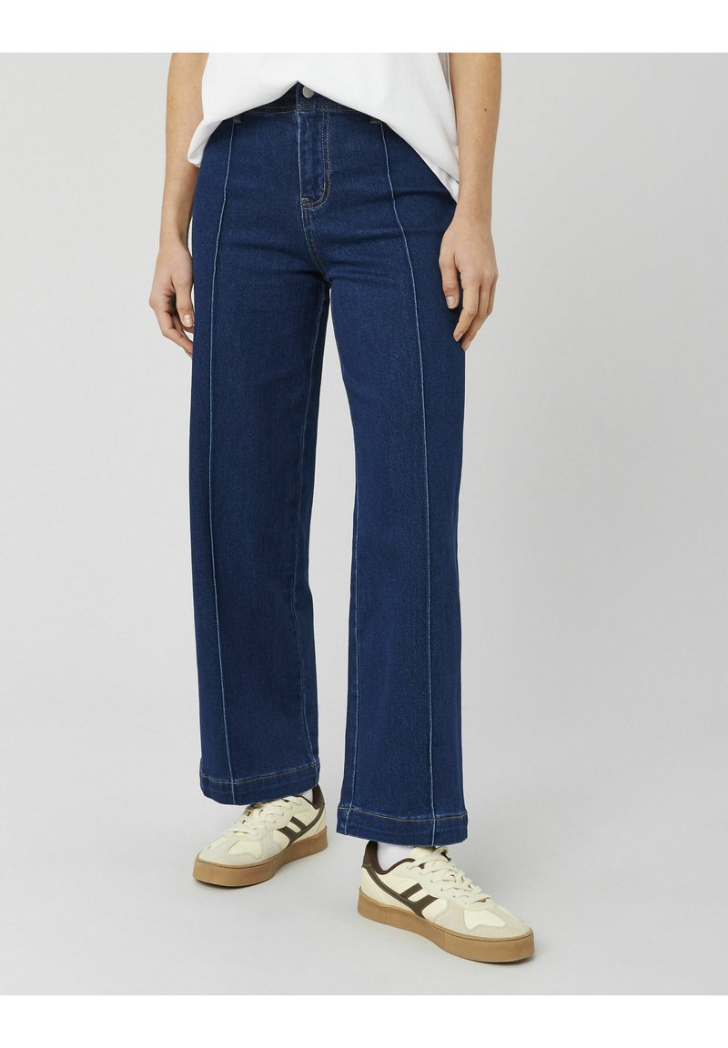 Jean bleu à jambes larges en denim, avec des surpiqûres contrastantes, une taille haute et une coupe droite, associé à des baskets crème.