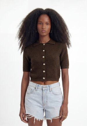 Žena s kučeravými vlasmi, nosiaca krátky hnedý zapínací crop top s krátkym rukávom a svetlomodré ošúchané šortky z džínsoviny, stojaca na bielom pozadí.