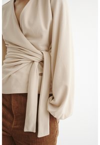 Top bege envolto com mangas longas e fluidas e um laço na cintura, feito de tecido macio. Combinado com calças de corduroy marrom texturizadas.