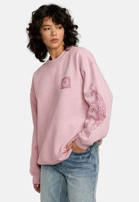 Sweatshirt oversized roz, fabricat dintr-un material moale. Prezintă un grafic mic cu soare și un model cu leopard pe mâneca stângă.