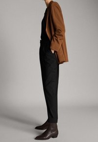 Braunes Sakko, schwarzes Oberteil und geschneiderte schwarze Hose. Strukturierte braune Stiefeletten mit vertikalen Akzenten. Schlanke Silhouette, minimalistisches Design.