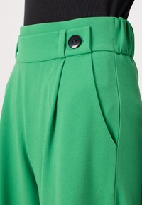 Pantalons tailleur verts en tissu lisse avec une taille haute, deux poches latérales et un détail boutonné à l'avant.