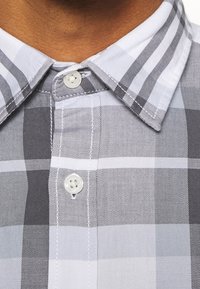 Camicia colletto a righe grigie e bianche con due bottoni bianchi visibili allacciati al collo e al petto.