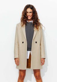 Manteau long beige au design ajusté, à double boutonnage à l'avant et boutons noirs, superposé sur un sweat-shirt gris et une chemise blanche.