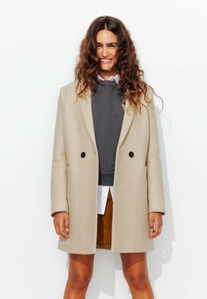 Manteau long beige au design ajusté, à double boutonnage à l'avant et boutons noirs, superposé sur un sweat-shirt gris et une chemise blanche.