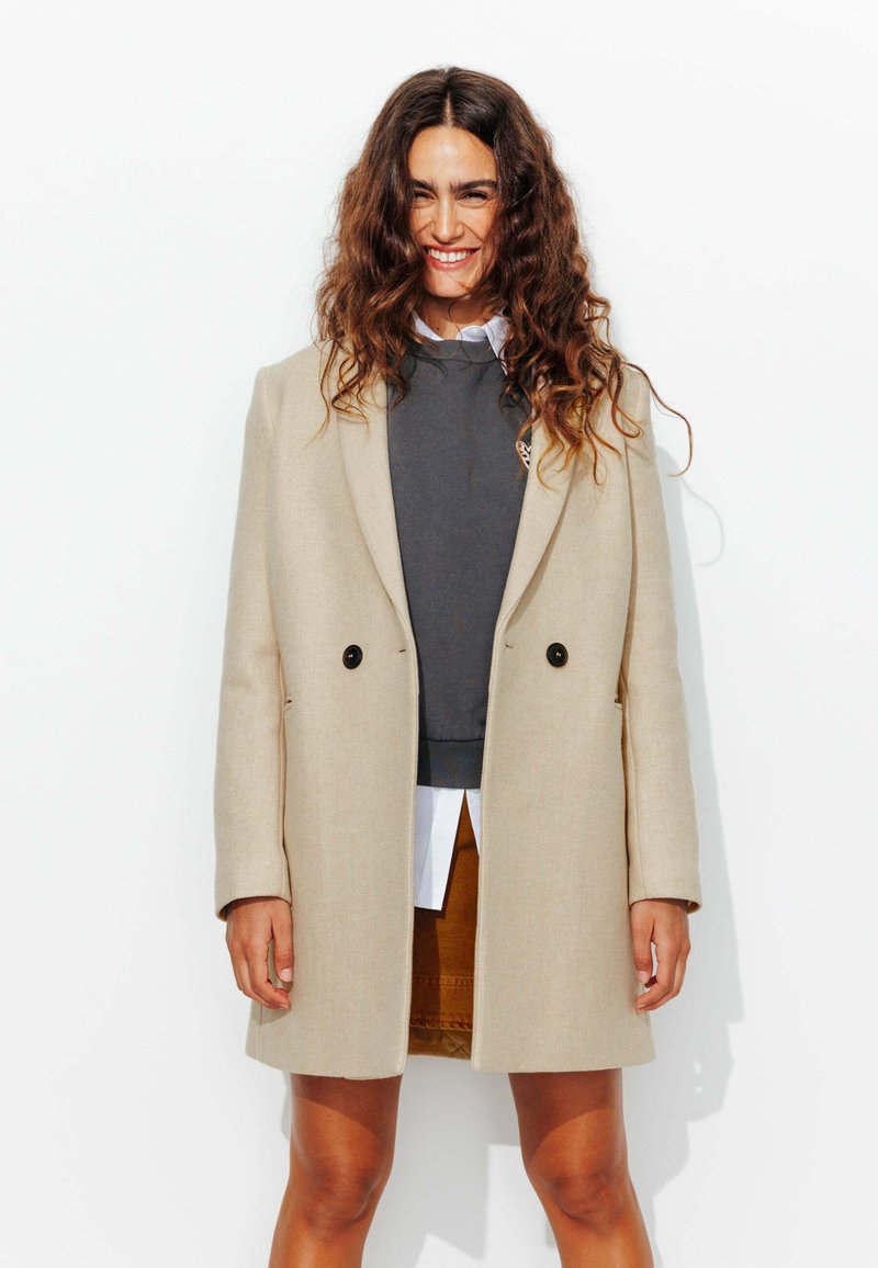 Manteau long beige au design ajusté, à double boutonnage à l'avant et boutons noirs, superposé sur un sweat-shirt gris et une chemise blanche.