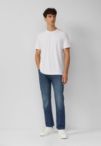 T-shirt bianco a maniche corte con colletto rotondo, abbinato a jeans in denim blu e sneaker bianche, in piedi contro uno sfondo neutro.