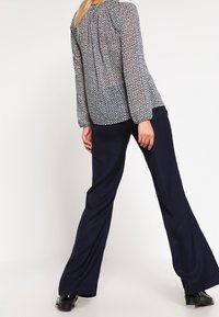 Blusa trasparente blu navy con un motivo geometrico, maniche lunghe e retro plissettato, abbinata a pantaloni larghi blu navy e scarpe nere.