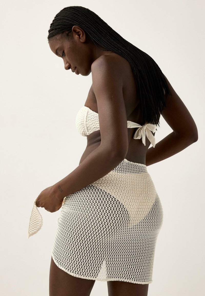 Kvinde med langt fletninghår iført hvid strikket bikinioverdel og matchende netnederdel, justerer nederdelen mod en ensfarvet baggrund.