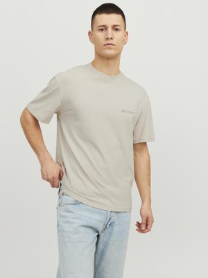 Beige short-sleeve t-shirt made of cotton. Crew neck design with small text detail on the left side. Paired with light blue denim jeans.
