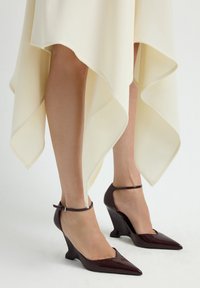 Zapatos de cuña burdeos brillantes con punta afilada y correa en el tobillo, combinados con un vestido de gasa de color crema con dobladillo asimétrico.