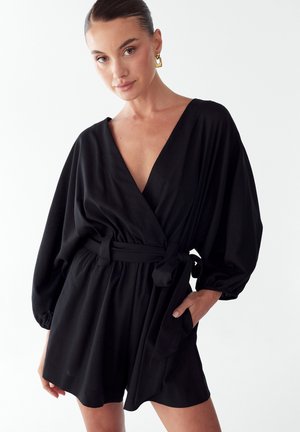 Žena s uhladenými vlasmi, ktorá nosí zlaté náušnice a čierny overal s opaskom a dlhými rukávmi, stojaca na bielom pozadí.