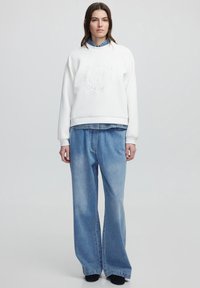 Sudadera de algodón blanca con logotipo en relieve, combinada con jeans de mezclilla azul claro de corte holgado, que presentan un tiro recto y un ligero desgaste.