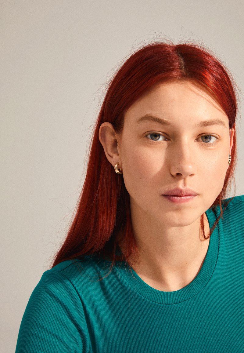 Türkisfarbenes geripptes Oberteil mit rundem Ausschnitt. Getragen mit kleinen goldenen Creolen. Lange rote Haare und Natur-Make-up. Neutraler Hintergrund.