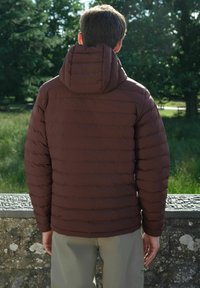 Hnedá puffa bunda s kapucňou, s prešívaným dizajnom a horizontálnymi pruhmi. Hladká textúra s voľným strihom. Nosená s svetlými nohavicami.