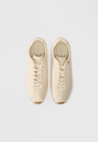 Baskets couleur crème en cuir lisse, avec un design minimaliste, un système de laçage et une semelle en caoutchouc avec un petit logo en accent.