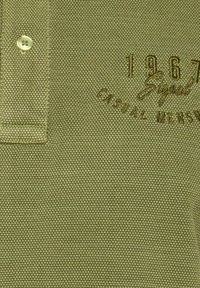 Pikepaita oliivinvihreänä, tekstuurikankaasta, jossa on kaulus, kaksi nappia ja brodéerattu '1967 Signal Casual Men's' logo.