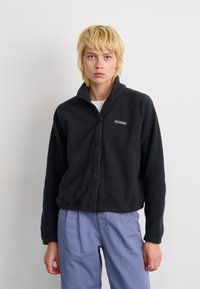 Black fleece jacket with a collared neckline, snap closure, and a left chest pocket featuring a logo. Worn with light blue pants.