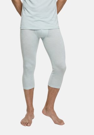 Man wearing light blue three-quarter length underwear and matching top, standing barefoot with hands behind back.