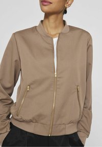 Femme portant une veste bombardier beige à fermeture éclair avec des zips dorés et un col, des poignets et un ourlet côtelés, superposée à un haut blanc et un pantalon noir.