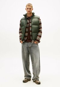 Grüne Steppweste über einem gestreiften Flanellhemd, kombiniert mit lockeren grauen Jeans und gemusterten Turnschuhen. Einfaches Design, funktioneller Stil.