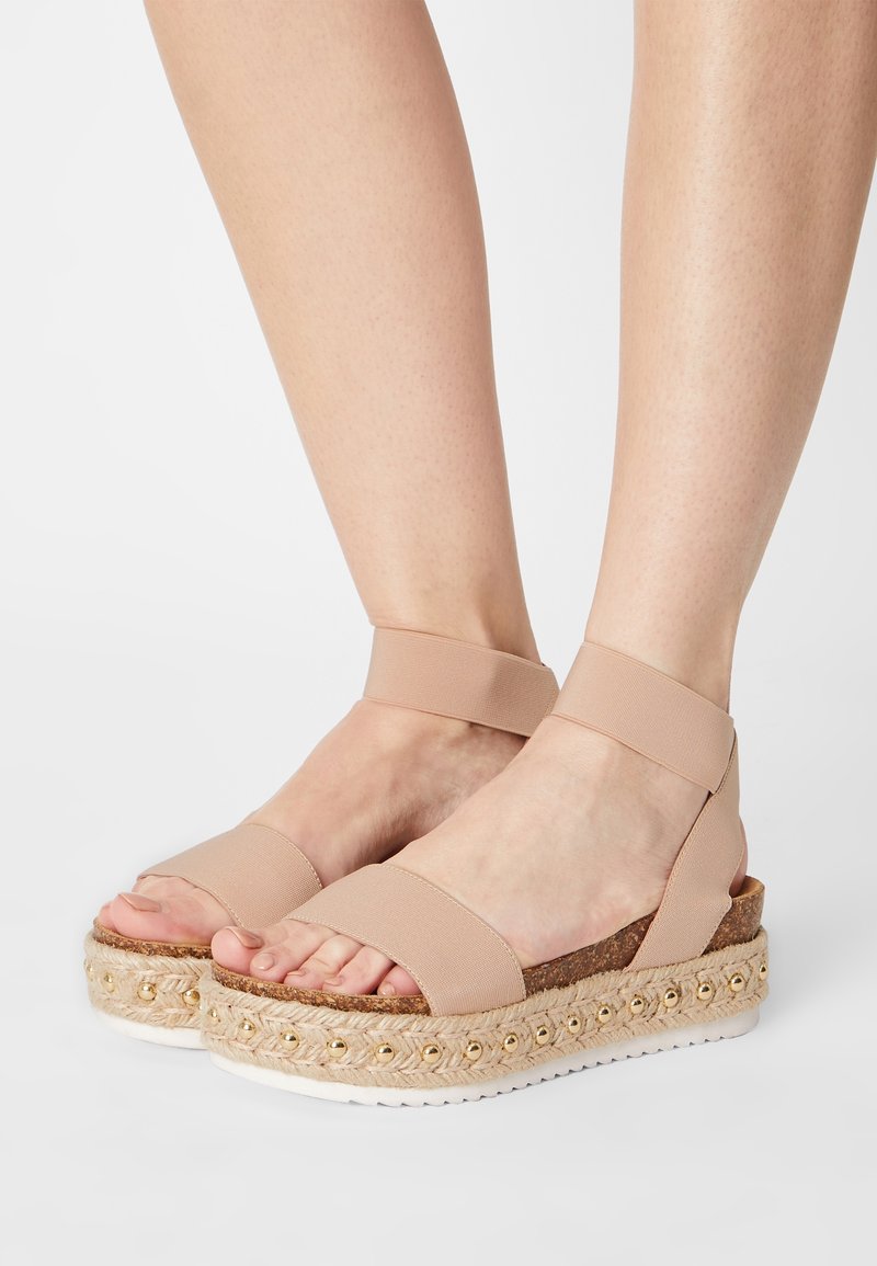 Pieds portant des sandales compensées beige avec des brides à la cheville et des clous dorés décoratifs sur des semelles en corde tressée sur fond blanc.