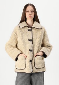 Beige nepbont jack met grote kraag, bruine afwerking en knoopsluitingen; heeft twee voorkeurszakken en een zachte, gestructureerde uitstraling.