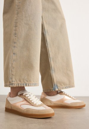 Stopy noszące beżowo-białe sneakersy Karl Lagerfeld z gumowymi podeszwami w połączeniu z szerokimi, jasnobrązowymi spodniami dżinsowymi.