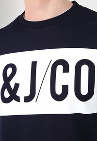 Marinblå crewneck sweatshirt med en vit horisontell bort med svart text "&/J/CO". Tillverkad av mjukt tyg med ribbat halsringning.