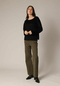 Vrouw staat in een fuzzy zwarte trui, olijfgroene wijde broek en zwarte haklaarzen tegen een effen achtergrond.