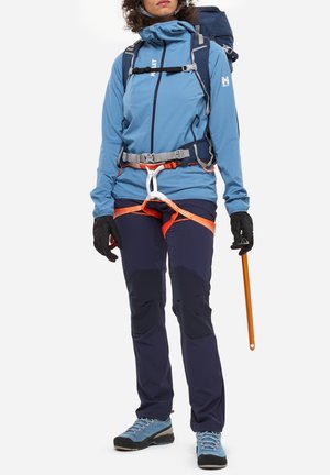Personne portant une veste bleue, un harnais d'escalade, des gants et un sac à dos, tenant un piolet orange, vêtue pour l'alpinisme.
