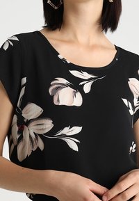 Blusa negra con flores y mangas cortas, que presenta grandes patrones florales en beige y blanco, un escote redondo y una tela ligera y texturizada.