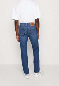 Jean en denim bleu avec une coupe droite, présentant une patch en cuir sur la taille et des poches arrière avec une couture distinctive.