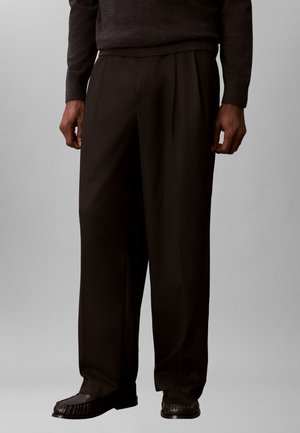 Uomo che indossa pantaloni neri larghi plissettati e scarpe slip-on nere texturate, in piedi davanti a uno sfondo grigio uniforme.