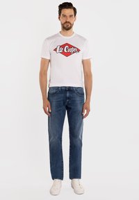 Mężczyzna ubrany w białą koszulkę z logo Lee Cooper, niebieskie dżinsy i białe trampki, stojący na tle jednolitego białego tła.