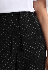 Pantalon noir à motifs de petits points blancs, doté d'une ceinture nouée à la taille et d'une texture de tissu lisse.