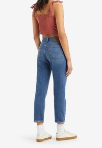 Femme portant un jean bleu taille haute, des baskets blanches, des chaussettes blanches et un crop top sans manches de couleur rouille avec des bretelles à nouer, debout de dos à la caméra.