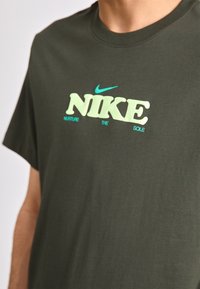 Mørkegrøn bomulds T-shirt med "NIKE" logo i lys gul og aqua, med sætningen "NURTURE THE SOLE" i mindre tekst nedenfor.