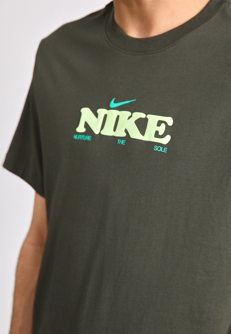 Ciemnozielona bawełniana koszulka z logo "NIKE" w jasnym żółtym i aqua, z frazą "NURTURE THE SOLE" w mniejszym rozmiarze poniżej.